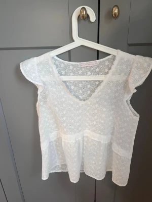 Vit blus från Version Feminin - Söt vit blus från Version Feminin i storlek S. Blusen har broderade blommor, v-ringning och volangdetaljer vid ärmarna. Tillverkad i lätt och luftig bomull, perfekt för varma dagar. Passar dig som gillar romantisk och feminin stil.