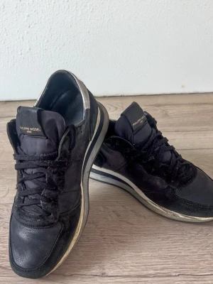 Svarta Philippe Model sneakers - Snygga svarta sneakers från Philippe Model med silvriga detaljer på hälen och sidan. Skorna har en klassisk låg siluett, svarta skosnören och en vit sula med svarta inslag. Materialmix av mocka och textil ger en cool och modern look.