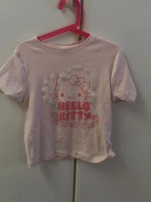 Ljusrosa Hello Kitty t-shirt - Söt ljusrosa t-shirt med Hello Kitty-tryck framtill och hjärtdetalj i bakgrunden. Klassisk rund halsringning och korta ärmar. Perfekt för dig som gillar gulliga motiv och vill ha en bekväm vardagstopp.