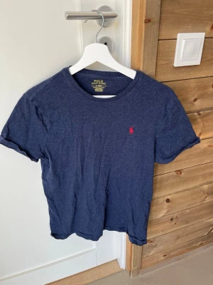 Marinblå t-shirt från Polo Ralph Lauren - Snygg marinblå t-shirt från Polo Ralph Lauren med röd broderad logga på bröstet. Modellen är custom slim fit och har klassisk rund hals och korta ärmar. Perfekt till jeans eller shorts för en clean och stilren look.
