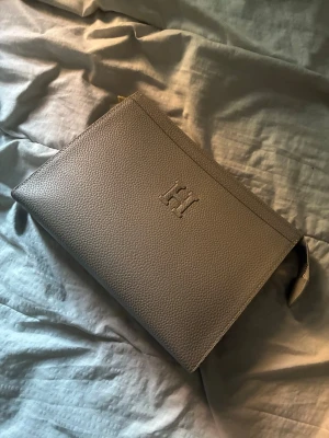 Beige kuvertväska i skinn - Stilren beige kuvertväska i grained skinn med diskret präglad logga på framsidan. Väskan har dragkedja upptill och en liten handledsrem. Perfekt för att bära det viktigaste och ger en clean look till din outfit.