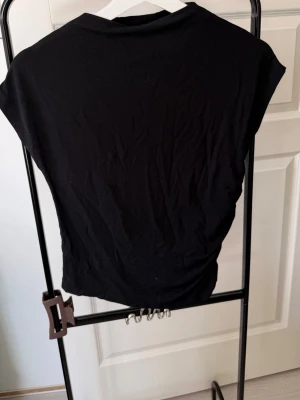 Svart topp med rynkad sida - En svart topp med korta ärmar och rynkade detaljer på sidorna. T-shirten har en enkel, stilren design och är tillverkad i ett mjukt material som känns bekvämt mot huden. Perfekt för dig som gillar minimalistisk stil.