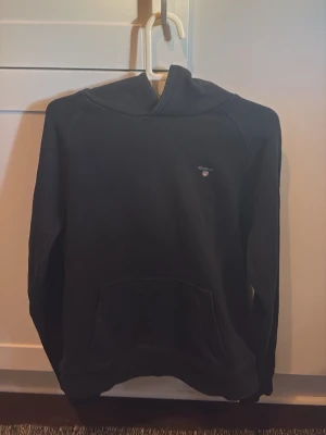 Svart hoodie från GANT - Snygg svart hoodie från GANT med liten broderad logga på bröstet. Klassisk känguruficka framtill och huva. Tillverkad i mjukt material som känns skönt mot huden. Perfekt för en avslappnad och stilren look.