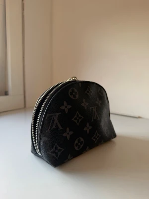 Svart monogram nessecär med dragkedja - Snygg svart nessecär i halvmåneform med silverfärgad dragkedja och klassiskt monogrammönster. Tillverkad i tåligt konstläder med stilrena detaljer. Perfekt för kort, mynt och småsaker.