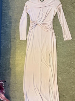 Ljusrosa långklänning med twistdetalj - Säljer en elegant ljusrosa långklänning med lång ärm och twistad detalj i midjan. Klänningen har en slits på sidan och är tillverkad i ett mjukt, stretchigt material som ger en snygg passform. Perfekt för dig som vill ha en stilren och modern look.