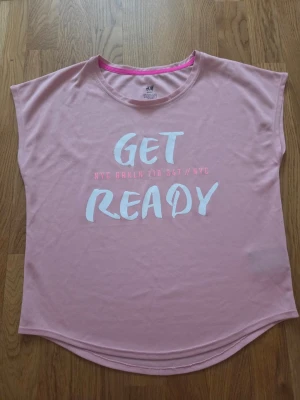 Rosa t-shirt med tryck från H&M - Ljusrosa t-shirt från H&M med vitt och rosa tryck på framsidan där det står 'GET READY'. T-shirten har rund halsringning, korta ärmar och är gjord i mjuk bomull. Perfekt för en avslappnad och trendig look.