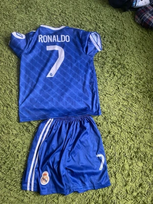Blå Real Madrid Ronaldo matchställ - Säljer ett blått Real Madrid matchställ med Ronaldo 7-tryck. Tröjan har vita detaljer, klubbmärke och 'Emirates Fly Better' på bröstet. Shortsen har vita ränder på sidorna och nummer 7 samt klubbmärke. Perfekt för fotbollsträning eller match.