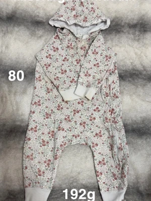 Blommig overall med huva, stl 80 - Söt overall med blommigt mönster i rosa och grönt på vit botten. Overallen har huva, långa ärmar med vita muddar och dragkedja längs sidan. Perfekt för mysiga dagar hemma eller ute. 🌸 Missfärgade muddar. Storlek 80