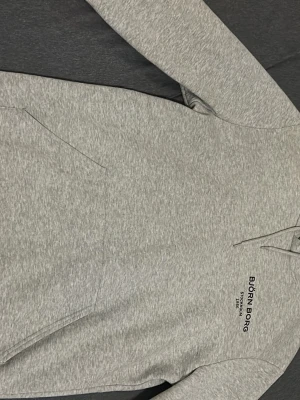 Ljusgrå hoodie från Björn Borg - Ljusgrå hoodie från Björn Borg med svart tryck på bröstet. Klassisk modell med huva och känguruficka framtill. Tillverkad i mjukt sweatshirtmaterial, perfekt för chill dagar eller träning. Snygg och enkel design som passar till det mesta.