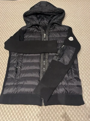 Svart pufferjacka med huva från Moncler - Snygg svart pufferjacka från Moncler med huva och ribbstickade ärmar. Jackan har dragkedja framtill, två fickor med knappar och en extra ficka med dragkedja på ärmen. Moncler-logga på vänster ärm. Perfekt för kalla vinterdagar.