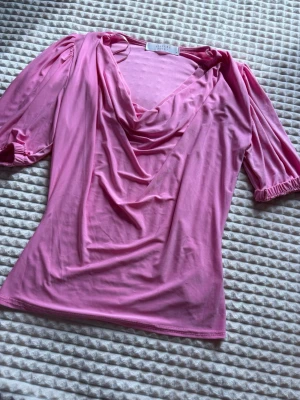 Rosa topp med draperad urringning - Säljer en rosa topp med draperad v-ringad urringning och puffiga korta ärmar med resår. Toppen har en mjuk och stretchig känsla, perfekt för dig som gillar en feminin och trendig stil. Materialet känns som polyester och toppen är figurnära.