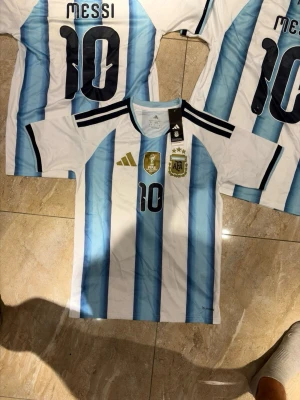 Argentina Messi matchtröja blåvit - Säljer en officiell Argentina matchtröja från Adidas med Messi och nummer 10 på ryggen. Tröjan har klassiska blå och vita ränder, svarta detaljer på axlarna och officiella klubbmärken framtill. Perfekt för dig som älskar fotboll och vill stötta ditt lag.