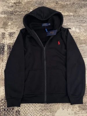 Svart hoodie från Polo Ralph Lauren - Snygg svart hoodie från Polo Ralph Lauren med dragkedja framtill och klassisk röd logga broderad på bröstet. Hoodien har två fickor fram, ribbade muddar och en mjuk insida. Perfekt för en chill och stilren look.