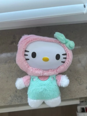 Hello Kitty mjukisdjur pastell - Söt Hello Kitty mjukisdocka med pastellfärger. Dockan har ett vitt ansikte, rosa huva med mintgrön rosett och mintgrön overall. Tillverkad i mjukt plyschmaterial och perfekt som dekoration eller samlarobjekt.