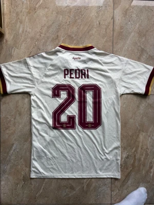 Vit Spanien fotbollströja Pedri #20 - Säljer en vit Spanien fotbollströja från Adidas med Pedri och nummer 20 på ryggen. Tröjan har vinröda detaljer, gula inslag på krage och ärmslut samt tre klassiska Adidas-ränder på axlarna. Perfekt för dig som älskar landslagsfotboll och vill stötta Spanien.