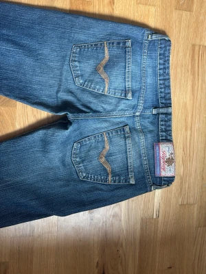 Jätte lowaist Blå bootcut jeans med broderade fickor - Säljer ett par blå bootcut jeans med snygga broderade detaljer på bakfickorna. Jeansen har klassisk femficksmodell, normal midja och är gjorda i slitstarkt denimtyg. Perfekta för en avslappnad och trendig look.