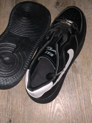 Svarta Nike Air Force 1 sneakers - Snygga svarta Nike Air Force 1 sneakers med vit swoosh och klassisk låg siluett. Skorna har svart ovandel i läder, perforerad tå och tjock svart sula. Tydlig Nike Air-logga på plösen och robusta svarta skosnören. Perfekta för en streetwear-look.