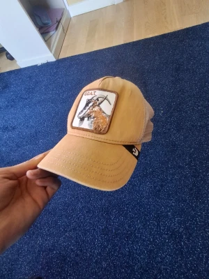 Goat keps hatstore - Säljer en beige truckerkeps med mesh på baksidan och ett broderat patch framtill med en get och texten 'GOAT'. Kepsen har böjd skärm och justerbar snapback. Perfekt för dig som gillar djurmotiv och streetwear. 