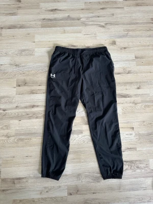Svarta mjukisbyxor från Under Armour - Säljer ett par svarta mjukisbyxor från Under Armour med resår i midjan och dragsko. Byxorna har en avslappnad passform, muddar vid bensluten och logga på vänster lår. Perfekta för chill eller träning.