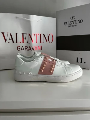 Valentino sneakers - Snygga vita sneakers med rosa band och ikoniska nitar i metall på sidorna. Skorna har en klassisk låg siluett, vita snören och detaljerad sula. Tillverkade i läder med exklusiv känsla och cool design som sticker ut.