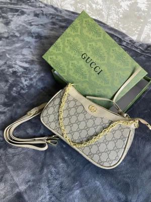 Beige axelremsväska från Gucci - Snygg beige handväska från Gucci med klassiskt GG-monogram i canvas och detaljer i ljusbeige läder. Väskan har en guldfärgad kedja som handtag och avtagbar axelrem. Perfekt för dig som gillar lyxiga accessoarer och tidlös design.