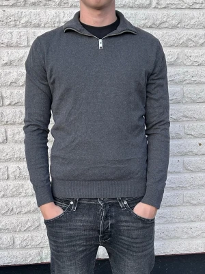 Mörkgrå tröja med halv dragkedja - Mörkgrå långärmad tröja med halv dragkedja i halsen och ribbade muddar vid ärmslut och nederkant. Tröjan har en stilren och enkel design, perfekt för lager på lager och funkar till många olika stilar. Storlek XS