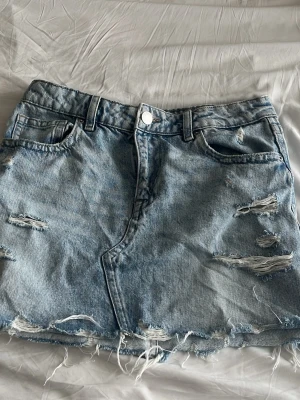 Ljusblå jeanskjol med slitningar - Säljer en kort ljusblå jeanskjol med slitna detaljer och rå kant. Kjolen har klassisk femficksmodell, bälteshällor och dragkedja med knapp framtill. Perfekt för dig som gillar en avslappnad och trendig stil.