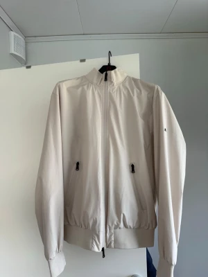 Beige vindjacka med dragkedja - Stilren beige vindjacka med hög krage och hel dragkedja framtill. Två fickor med dragkedja och ribbade muddar vid ärmslut och nederkant. Lätt material som passar perfekt för blåsiga dagar. Jackan har några fläckar som inte gått bort, det är små svarta oljefläckar och en annan fläck (fler bilder kan skickas vid efterfrågan. Säljes billigare pga detta. Nypris 1800 förra året.