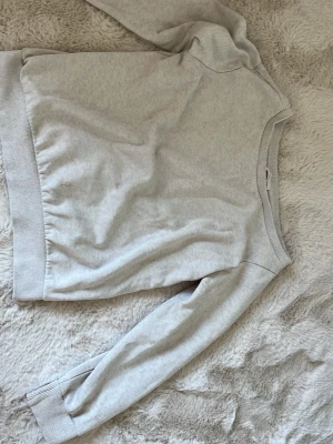 Ljusgrå basic offshoulder tröja - En enkel och stilren ljusgrå offshoulder tröja med långa ärmar och ribbade muddar vid ärmslut och nederkant. Tillverkad i mjukt sweatshirtmaterial som känns skönt mot huden. Perfekt för en avslappnad look. Den har 2 st fläckar (syns på bild 2, men man ser dem knappast)! 