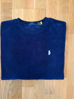 Polo Ralph Lauren T-Shirt - Polo Ralph Lauren T-Shirt i mörkblå färg med den klassiska Polo-hästen broderad på bröstet. Den är mycket fint skick och är bra omhändertagen. Tröjan är i storlek S och har en ganska avslappnad passform som inte sitter för tajt men ändå inte för löst. OBS! Tröjan har en mjuk yta med en frottéliknande känsla och är tillverkad i 88 % bomull och 12 % polyester. Om du har frågor så ställ dem gärna.
