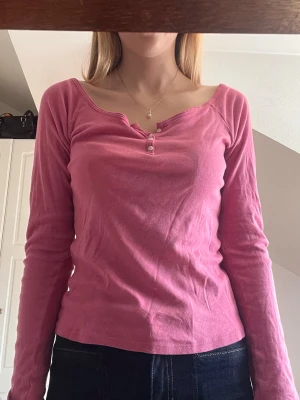 Rosa långärmad topp med knappar - Säljer en rosa långärmad topp med offshoulder-design och små knappar framtill vid halsringningen. Toppen har en smickrande passform och är gjord i mjukt bomullsmaterial. Perfekt för dig som vill ha en enkel men snygg topp till garderoben. 100% bomull