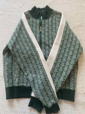 Grön mönstrad Lacoste hoodie med dragkedja - Snygg grön hoodie med vitt geometriskt mönster och dragkedja framtill. Tröjan har ribbade muddar vid ärmslut och nederkant samt hög krage. Vita ränder längs ärmarna ger en sportig look. Perfekt för dig som gillar streetwear och unika detaljer.