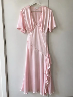 Ljusrosa omlottklänning i satin - Superfin ljusrosa långklänning i satin med omlottmodell och v-ringning. Klänningen har korta ärmar och en snygg volangdetalj längs sidan. Perfekt för dig som vill ha en romantisk och elegant look.
