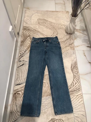 Blå raka jeans med hög midja - Klassiska blå jeans med rak passform och hög midja. Jeansen har fem fickor, knappgylf och subtila kopparfärgade nitar. Materialet är kraftig denim i 100% bomull. Perfekta för en avslappnad och tidlös stil.
