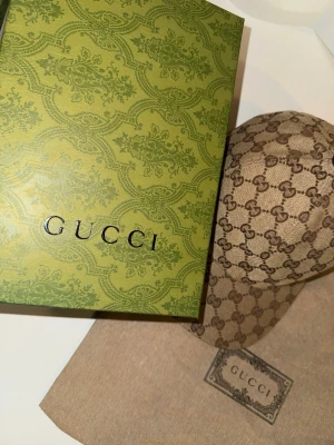 Gucci beige keps med GG-mönster - Säljer en beige keps från Gucci med det klassiska GG-monogrammet över hela kepsen. På sidan finns en grön och röd rand som ger den en ikonisk look. Kepsen är tillverkad i canvasmaterial och har en böjd skärm. Perfekt för dig som vill ha en lyxig och trendig accessoar.