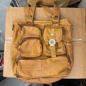 Brun handväska från Diesel - Cool brun handväska från Diesel i fejkskinn med flera praktiska ytterfack och detaljer i guld. Väskan har både handtag och axelremmar, samt en unik dekorativ knapp på framsidan. Perfekt för dig som gillar streetstyle och vill ha plats för allt. Nypris är 1799kr.