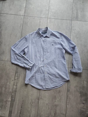 Blårandig skjorta från Ralph Lauren - Klassisk blårandig skjorta från Ralph Lauren med lång ärm och knappar framtill. Skjortan har en liten broderad logga på bröstet och är tillverkad i bomull. Perfekt för en stilren och avslappnad look.