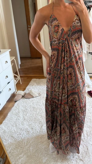 Mönstrad långklänning med halterneck - Säljer en bohemisk långklänning med färgstarkt paisleymönster i rött, beige och svart. Klänningen har halterneck med knyt i nacken, djup v-ringning och öppen rygg. Luftig passform och volang nedtill, perfekt för varma dagar.