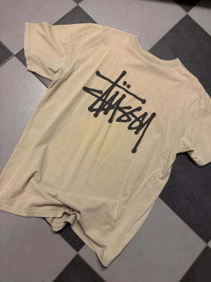 Beige Stüssy t-shirt med logga - Säljer en beige t-shirt från Stüssy med stor svart logga på ryggen. Klassisk passform och rund halsringning. Perfekt för dig som gillar streetwear och vill ha en enkel men snygg tee i garderoben.