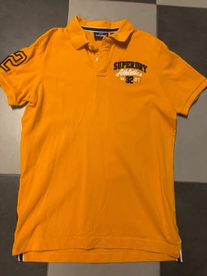 Gul pikétröja från Superdry - Säljer en gul pikétröja från Superdry med broderad text på bröstet och siffran 2 på ärmen. Tröjan har klassisk krage, korta ärmar och knappar vid halsen. Perfekt för dig som gillar sportig och avslappnad stil.
