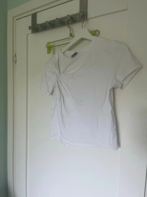 Vit t-shirt med twistad detalj - Säljer en enkel vit t-shirt med korta ärmar och en twistad detalj vid axeln. T-shirten har en klassisk passform och är gjord i mjuk bomull, perfekt till alla dina outfits.