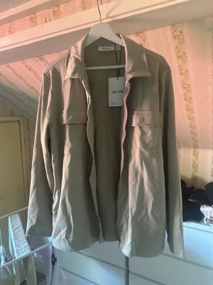 Beige overshirt från Only & Sons - Stilren beige overshirt från Only & Sons med klassisk krage och två bröstfickor med lock. Overshirten har långa ärmar och är tillverkad i ett mjukt tyg som ger en avslappnad look. Perfekt att bära öppen över en t-shirt. Bara testad och prislappen sitter kvar. Finns även sidofickor vid höften. Hör av er för mer info, pris är inte hugget i sten men skambudare behöver inte höra av sig