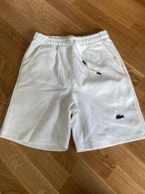 Vita shorts med snörning från Lacoste - Snygga vita shorts från Lacoste med elastisk midja och snörning. Klassisk krokodil-logga på vänster ben och två sidofickor. Perfekta för sommaren och tillverkade i mjuk bomull för maximal komfort.