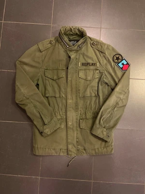 Olivgrön fieldjacket  från Replay - Snygg olivgrön fieldjacket från Replay med fyra fickor framtill, axelklaffar och märke på axeln. Jackan har ett snöre i vilket innebär att man kan dra åt jackan för att få den tightare. Jackan är perfekt nu till hösten, men ochså till våren eller sommaren. Modellen är 185 på bilden. Skriv gärna om ni har frågor!😃🙌🙌🙌