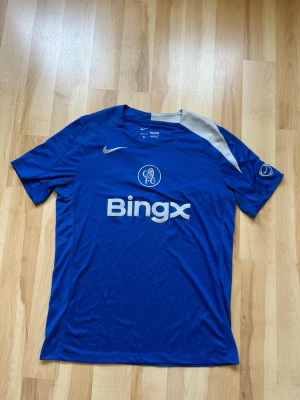 Blå Chelsea FC matchtröja Nike - Säljer en blå Chelsea FC matchtröja från Nike med Bingx-logga på bröstet och klubbmärke framtill. Tröjan har korta ärmar, rund hals och en beige detalj vid axeln. Tillverkad i lätt och ventilerande material, perfekt för fotboll eller träning.