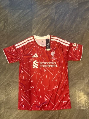 Röd Liverpool matchtröja Adidas 2026/2027 - Säljer en röd Liverpool FC matchtröja från Adidas med vita detaljer och grafiskt mönster. Tröjan har korta ärmar, rund hals och klubbmärke samt sponsortryck på bröstet. Perfekt för dig som älskar fotboll och vill stötta LFC.