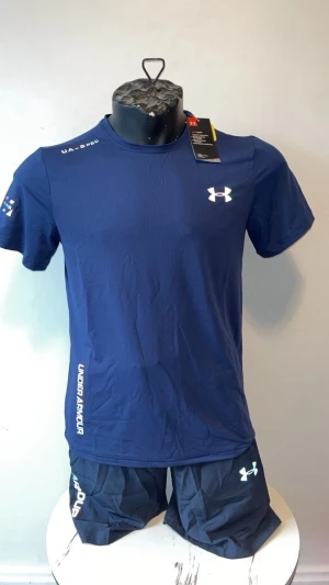 Blått träningsset från Under Armour - Snyggt träningsset från Under Armour i marinblått. Setet består av en kortärmad t-shirt och matchande shorts, båda med vita logotyper och tryck. Tillverkat i ett lätt och snabbtorkande material som passar perfekt för gymmet eller löpning.