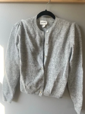 Grå kofta från Monki - Mysig grå kofta från Monki i storlek XS. Koftan har rund halsringning, ribbade muddar och knappar framtill. Perfekt att slänga över en t-shirt eller linne. Materialet är mjukt och ger en avslappnad vibe.
