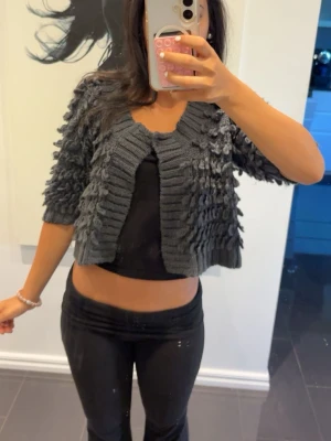 Mörkgrå kort stickad kofta/ bolero - Kortärmad, mörkgrå stickad kofta med strukturstickade detaljer och öppen framtill. Koftan har en chunky känsla med bred ribbstickning vid kanterna och ett coolt, puffigt mönster över hela plagget. Perfekt att slänga över en topp för en trendig lager-på-lager-look. passar S