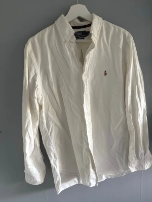 Vit skjorta från Polo Ralph Lauren - Klassisk vit skjorta från Polo Ralph Lauren med broderad logga på bröstet. Skjortan har lång ärm, knappar framtill och en tidlös design som passar till många olika stilar.